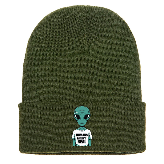 Olive green acrylic beanie with a cartoon alien wearing a white T-shirt that says “Humans Aren’t Real” in bold black letters.